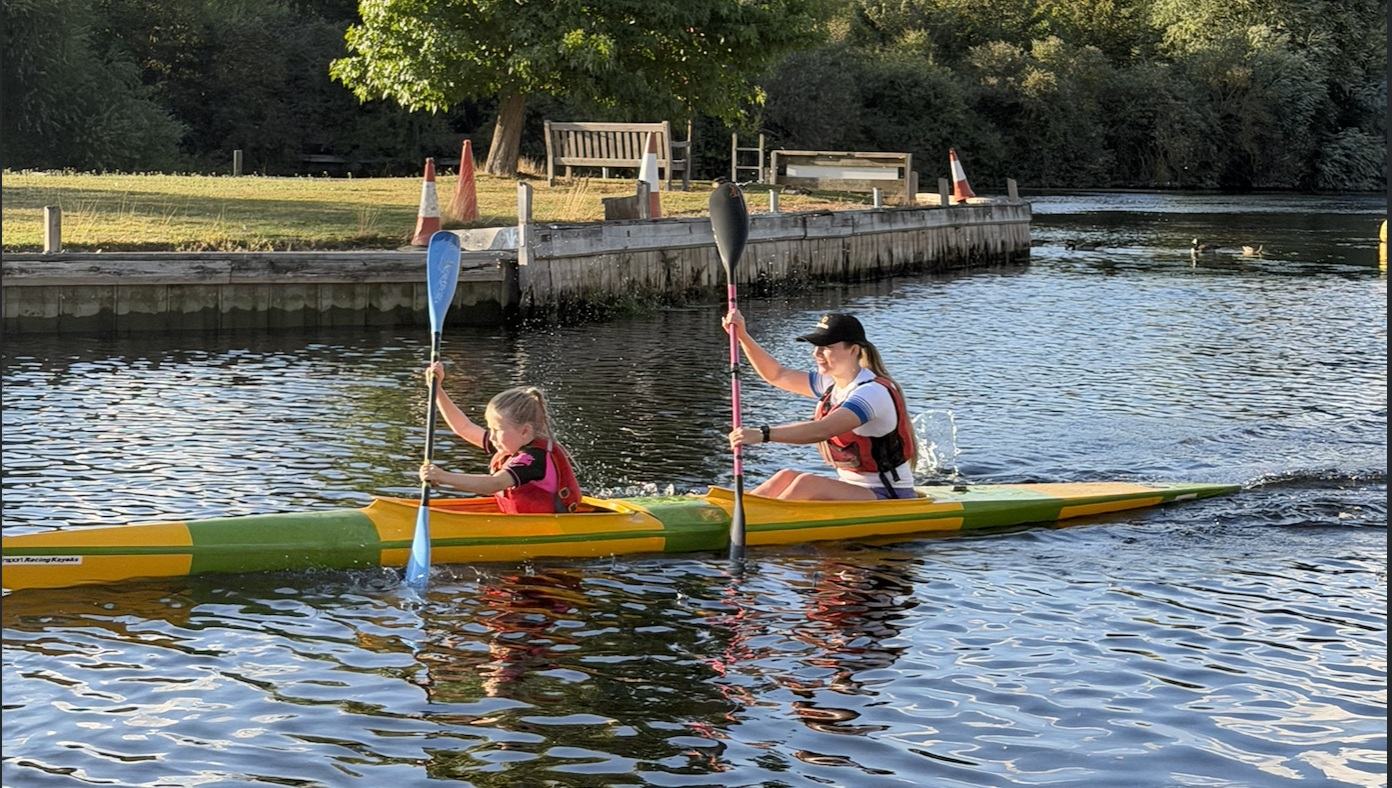 A coach and a young paddler training together in a K2 on flat water at the end of the day.